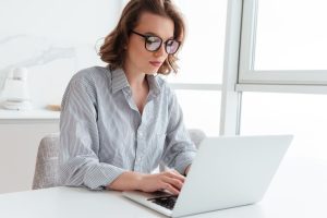 A imagem mostra uma mulher usando óculos e trabalhando em um laptop, concentrada no que está fazendo. Ela está em um ambiente claro e bem iluminado, com janelas grandes ao fundo, sugerindo que o local recebe muita luz natural. Sua postura é focada e parece estar envolvida em uma tarefa importante. O cenário é minimalista, com uma mesa branca e poucos objetos visíveis, o que reforça um clima de simplicidade e produtividade. Essa imagem é frequentemente associada a ambientes de trabalho remoto, estudos ou momentos de concentração em tarefas online.