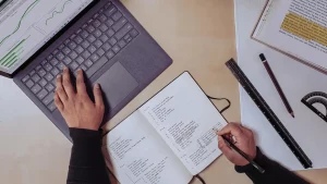 A imagem mostra uma vista aérea das mãos de uma pessoa trabalhando em uma mesa. Uma das mãos está sobre o teclado de um laptop, que exibe gráficos de dados na tela, sugerindo atividades de análise ou estudo. A outra mão está escrevendo em um caderno aberto, que contém anotações organizadas em listas. Ao redor do laptop e do caderno, há vários itens como uma régua, um transferidor, uma caneta e um livro aberto com partes destacadas em amarelo, indicando estudo ou planejamento. A cena transmite uma sensação de produtividade, planejamento e foco.