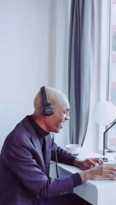 Um homem com cabelo platinado usa um terno escuro e fones de ouvido enquanto sorri, digitando em um laptop. Ele está sentado em uma mesa iluminada, com uma janela ao fundo e uma luminária ao lado, criando um ambiente moderno e acolhedor.