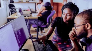 Duas pessoas colaborando em um ambiente de trabalho, com um computador de mesa mostrando código na tela, enquanto outros colegas trabalham em laptops ao fundo.
